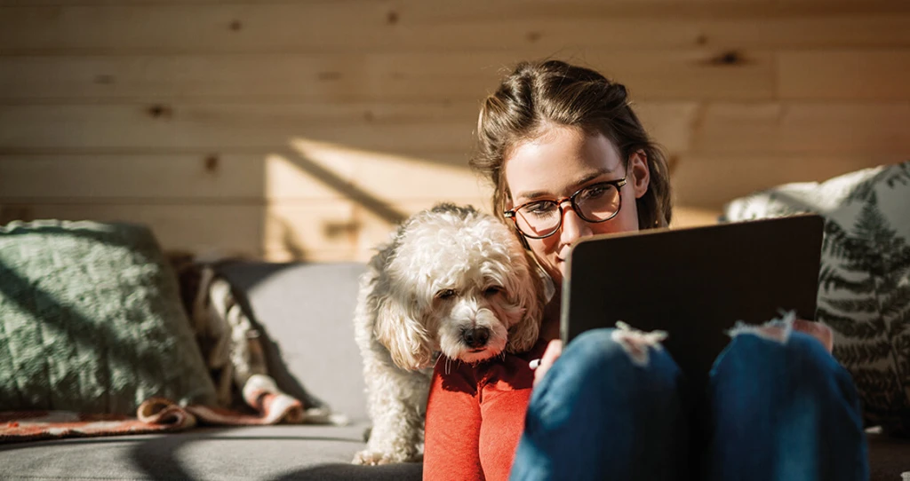Eine Frau am Tablet, ihr Hund schaut ihr über die Schulter. Im Hintergrund ein gemütliches Wohnzimmer mit Holzelementen.