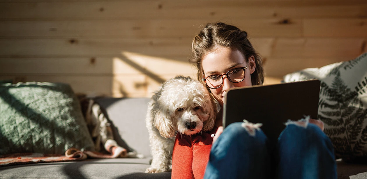 Eine Frau am Tablet, ihr Hund schaut ihr über die Schulter. Im Hintergrund ein gemütliches Wohnzimmer mit Holzelementen.
