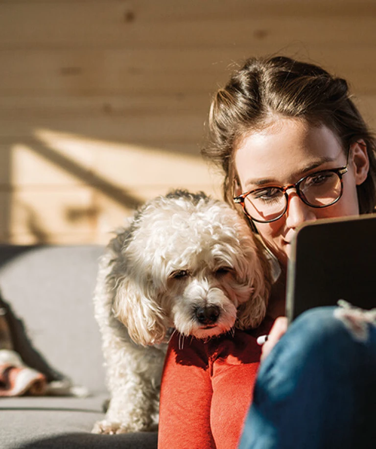 Eine Frau am Tablet, ihr Hund schaut ihr über die Schulter. Im Hintergrund ein gemütliches Wohnzimmer mit Holzelementen.