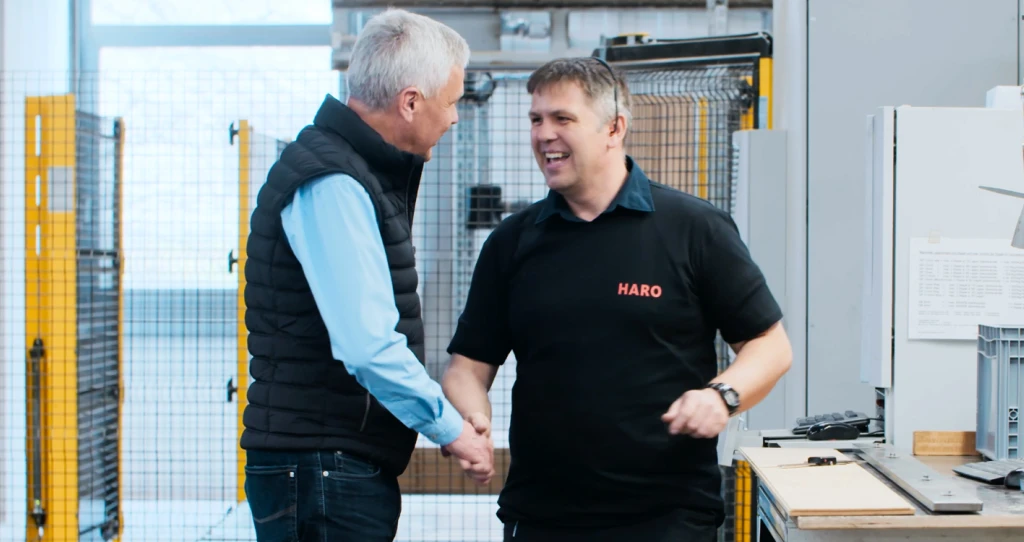 Zwei Männer stehen in einer Produktionshalle und schütteln sich die Hand – symbolisch für Zusammenarbeit, Vertrauen und den menschlichen Aspekt moderner Fertigung.