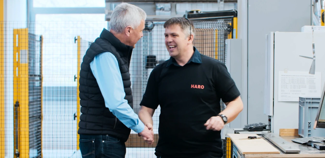Zwei Männer stehen in einer Produktionshalle und schütteln sich die Hand – symbolisch für Zusammenarbeit, Vertrauen und den menschlichen Aspekt moderner Fertigung.