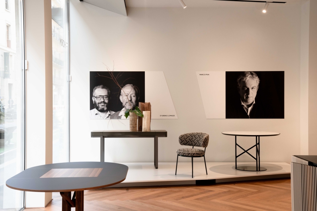 La imagen muestra un showroom luminoso con parquet HARO oscuro en espiga como contraste. En la pared cuelgan imágenes de reconocidos diseñadores de muebles.